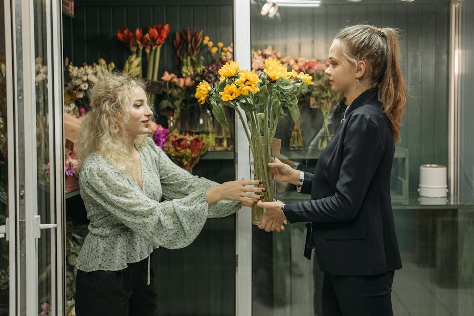 A florist customer and staff member exchange a bright yellow sunflower bouquet wrapped in clear plastic in a flower shop. The customer, a young woman with curly blonde hair wearing a light-colored, pa