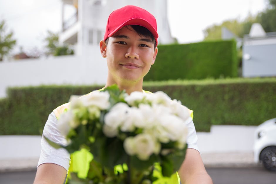 A delivery person wearing a red cap and a high-visibility vest stands outdoors on a residential street, holding a vibrant floral bouquet with white and green flowers. The bouquet appears freshly arran