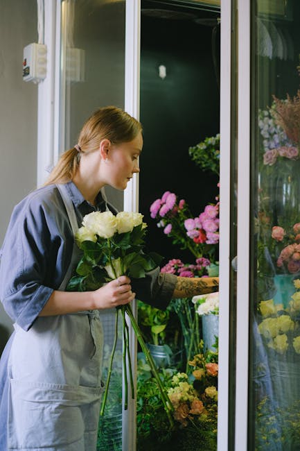 A young woman with fair skin and reddish-brown hair tied back, dressed in a light gray work jacket, is holding a bouquet of fresh white roses with green foliage, inspecting them closely inside a flowe