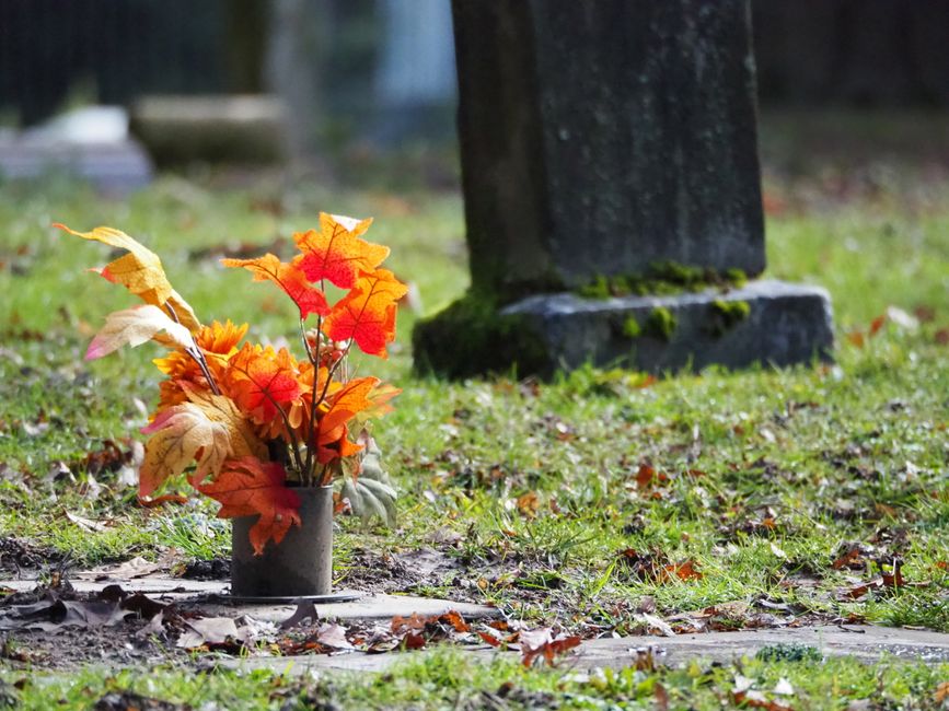A floral arrangement of artificial autumn leaves and flowers in shades of orange, red, yellow, and cream, arranged in a small, cylindrical grey vase. The bouquet is placed on a grave in a cemetery, wi