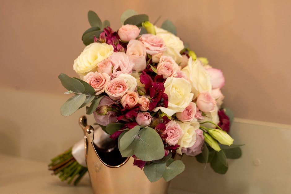 A floral bouquet featuring an arrangement of fresh white roses, pale pink spray roses, and deep burgundy blossoms, complemented by dusty green eucalyptus leaves. The bouquet is tightly clustered with