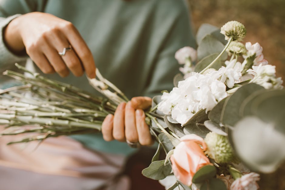 A florist from floristkingstonvale.co.uk is carefully arranging a floral bouquet featuring white hydrangeas, soft pink roses, and green foliage, with the stems wrapped in brown kraft paper. The floris