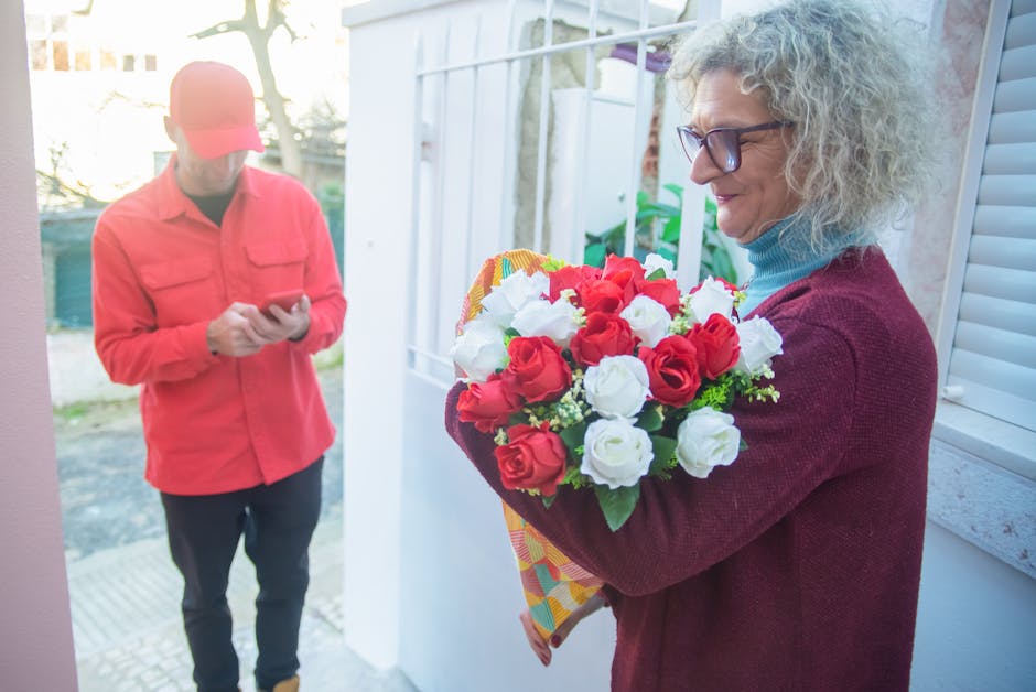 A woman with curly gray hair, wearing glasses and a burgundy sweater over a light blue turtleneck, is holding a bouquet of fresh red and white roses arranged with green foliage, wrapped in colorful pa