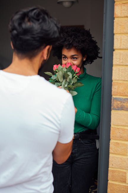 A woman with natural curly black hair, wearing a green long-sleeved top, stands at her doorway holding a bouquet of fresh pink roses with green foliage, ready for flower delivery. The bouquet is wrapp