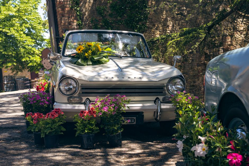 A vintage white car decorated with a large bouquet of yellow sunflowers and green foliage positioned on its hood, accompanied by an array of colorful potted flowers including pink, purple, and white b