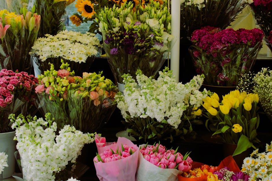 A vibrant display of freshly cut flowers arranged in bouquets and vases at a florist shop. The arrangement includes white stock, pink tulips wrapped in pink paper, yellow tulips, purple statice, white