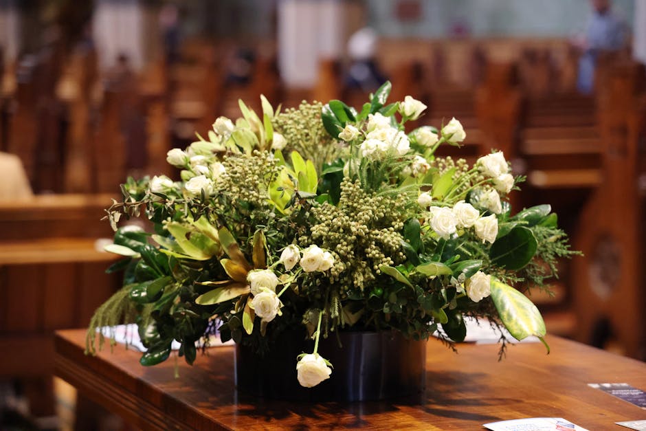 A lush floral arrangement featuring predominantly white flowers, including roses and small filler flowers, complemented by green foliage with broad and slender leaves. The bouquet is arranged in a bla