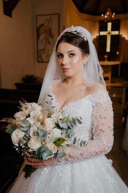 A bride inside a church, holding a large floral bouquet featuring white and blush roses, eucalyptus leaves, and other soft, pastel-hued flowers arranged in a cascading style. She is wearing an elegant