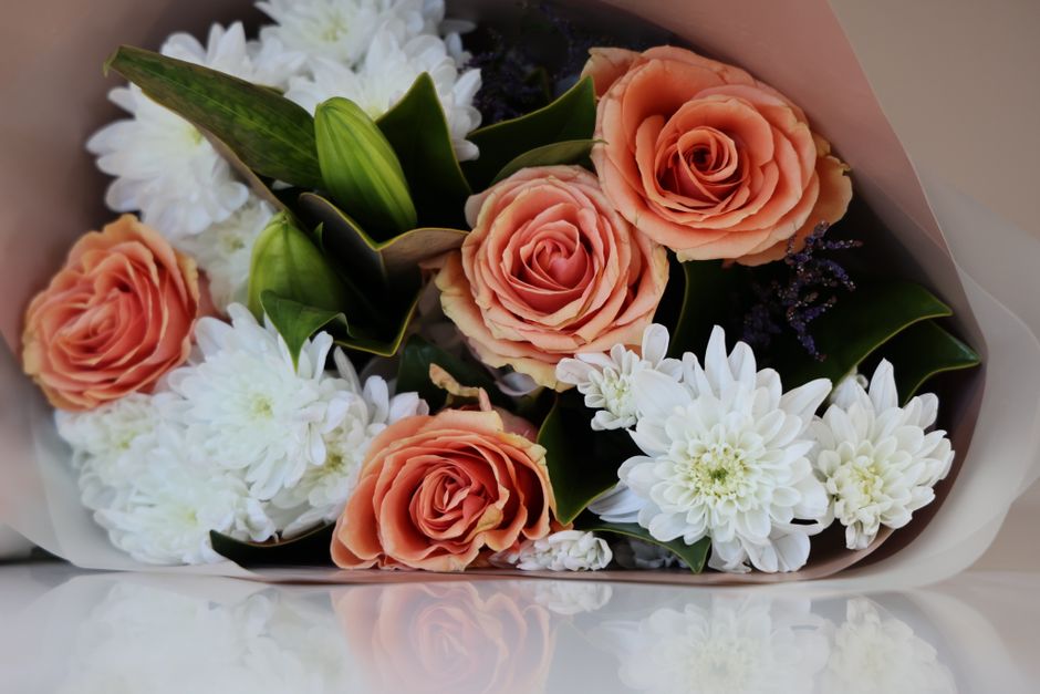 A floral arrangement featuring several peach-colored roses with gently unfurling petals, complemented by white chrysanthemums with layered petals, and accented with dark green leaves and small cluster