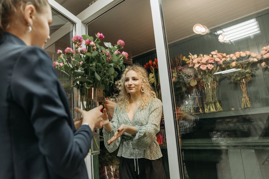 A young woman with curly blonde hair is inside a flower shop near the glass door, smiling as she receives a bouquet of fresh pink roses with green foliage from a florist. The florist, partially visibl