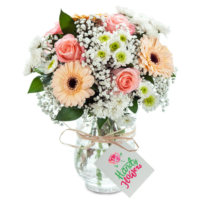 Bathed in natural daylight, this elegant bouquet, inspired by the green heart of Kingston Vale, is arranged with a florist's precision and sensitivity to colour. Several lush, pastel peach-pink roses unfurl at the centre, surrounded by two striking peach-hued gerberas whose wide, symmetrical petals and dark centres offer a cheerful pop. Wispy sprays of baby's breath filter light through the arrangement, lending an ethereal quality reminiscent of early morning mist over Wimbledon Common. Crisp white chrysanthemums with fresh lime centres dot the bouquet, infusing it with life and a nod to classic pick-your-own experiences at local gardens. Deep green eucalyptus, carefully placed for balance, provides natural architecture and a subtle aroma. The flowers are arranged in a clear vase that allows a full view of the submerged stems, grounded by a swath of rustic twine tied at the neck. A blank rectangular gift tag dangles from the bow, ready to carry messages for any occasion-ideal for thoughtful gifts or celebrations within Kingston Vale. Expert craft and local pride shine in every detail, making this bouquet suitable for home, work, or any gathering of loved ones.